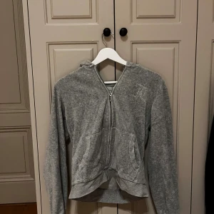 Grå Juicy Couture hoodie med strass - Grå hoodie från Juicy Couture med strassdetaljer på bröstet och stor Juicy Couture-logga i strass på ryggen. Tröjan har dragkedja framtill, huva och två fickor. Mjuk och skön i bomullsmix, perfekt för chill dagar. Klassisk och snygg streetstil.