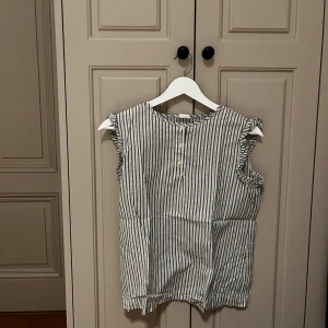 Randig blus från Pompdelux - Säljer en söt blus från Pompdelux i storlek 164. Den är vit med smala blå ränder, har volangdetaljer vid ärmhålen och knappar framtill. Blusen är ärmlös och gjord i mjuk bomull, perfekt för vår och sommar. Fräsch och stilren look!