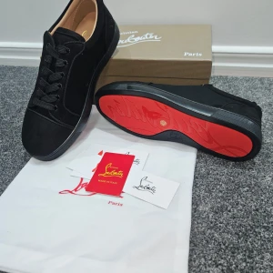 Svarta sneakers från Christian Louboutin - Snygga svarta sneakers från Christian Louboutin med klassisk röd sula och lyxig mocka på ovansidan. Skorna har rund tå, platt sula och svart snörning. Insidan är beige med guldfärgad logga. Perfekt för dig som vill sticka ut med exklusiv stil.