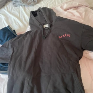 Svart hoodie från YOURTURN med tryck - Svart hoodie från YOURTURN med huva och känguruficka. På bröstet står det 'artist' i rött och på ryggen 'I lied that I'm an artist'. Mjuk insida och klassisk passform.