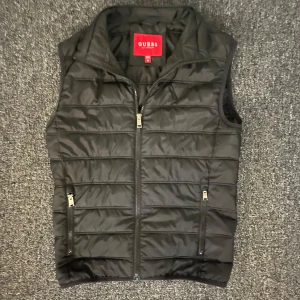 Svart dunväst från Guess  - Snygg svart dunväst från Guess i storlek XS. Västen har en bra design, dragkedja framtill och två fickor med dragkedja. Perfekt att slänga över en hoodie eller tröja när det är kyligt ute. Passar inte mig längre, sitter som en S.