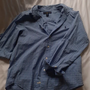 Blårutig skjorta från Ralph Lauren - Klassisk blårutig skjorta från Ralph Lauren med knappar framtill och broderad logga på bröstet. Skjortan har lång ärm, krage och är tillverkad i mjuk bomull. Perfekt för en avslappnad men ändå stilren look. Priset är ej hugget i sten!