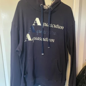 Mörkblå hoodie från Aquascutum - Snygg mörkblå hoodie från Aquascutum med stor logga framtill i vitt och blått. Huva med rutigt foder och dragsnören, samt klassisk känguruficka. Perfekt för dig som gillar streetwear med brittisk touch. Eller Ultras stilen. Passar även L