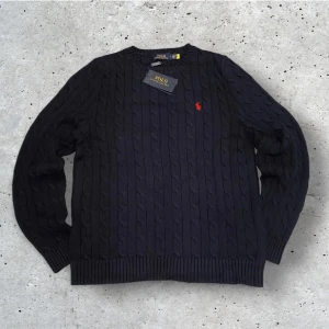 Ralph Lauren Stickad Sweater - Kabelstickad mörkblå tröja från Polo Ralph Lauren med klassisk röd broderad logga på bröstet. Perfekt inför en kall vinter!