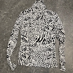 Zebramönstrad polotröja från H&M - Säljer en svartvit polotröja från H&M i storlek S. Tröjan har ett coolt zebramönster, lång ärm och hög polokrage. Tillverkad i mjuk bomullsmix som är skön mot huden. Perfekt för dig som vill sticka ut med ett djurmönster i garderoben.