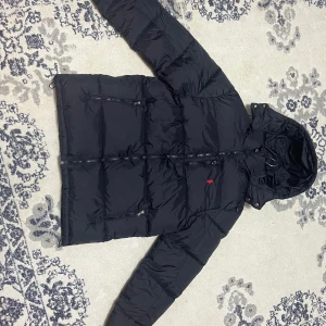 Svart dunjacka från Ralph Lauren - Svart dunjacka från Ralph Lauren med huva och flera dragkedjeförsedda fickor. Jackan har klassisk pufferstil och är dekorerad med den ikoniska röda Polo-loggan på bröstet. Perfekt för kalla dagar och har en snygg, sportig look. Helt oanvänd.