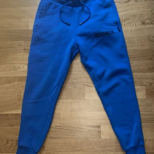 Blå mjukisbyxor från Hoodrich - Snygga blå mjukisbyxor från Hoodrich med svart logga på låret. Byxorna har resår och snörning i midjan samt muddar vid bensluten. Perfekta för en avslappnad stil och riktigt sköna att ha på sig.