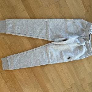 Grå mjukisbyxor från U.S. Polo Assn. - Grå mjukisbyxor från U.S. Polo Assn. med broderad logga på benet. Byxorna har snörning i midjan, ribbade muddar vid bensluten och är gjorda i en mjuk bomullsblandning. Perfekta för chill eller träning.