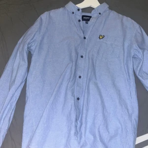 Blå skjorta från Lyle & Scott Junior - Snygg ljusblå skjorta från Lyle & Scott Junior med klassisk button down-krage och svarta knappar. Bröstficka med den ikoniska gula örnloggan. Skjortan är långärmad och i mjuk bomull, perfekt för en clean och stilren look.