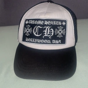 Chrome Hearts truckerkeps svart/vit - Säljer en stilren truckerkeps från Chrome Hearts med svart skärm och vit frontpanel. Kepsen har ett stort broderat märke framtill med texten 'Chrome Hearts Hollywood, USA' och ikoniska korsdetaljer. Baksidan är i svart mesh för extra ventilation.
