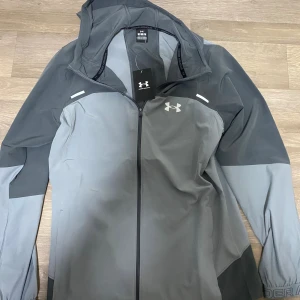 Grå vindjacka från Under Armour M - Snygg grå vindjacka från Under Armour med huva och hel dragkedja. Jackan har två olika grå nyanser, logga på bröstet och stor text på ryggen. Perfekt för träning eller när du vill ha en sportig look. Lätt och smidig modell med långa ärmar.