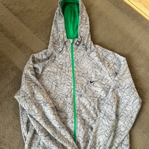 Nike vindjacka med grafiskt mönster - Säljer en grå Nike tröja med svart grafiskt mönster och grön insida i huvan. Jackan har hel dragkedja i grönt, två fickor med dragkedja och huva med snörning. Perfekt för dig som gillar sportig stil och snygga detaljer.Allt är bra med tröjan 