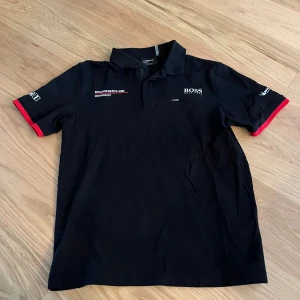 Svart Porsche x BOSS pikétröja M - HELT NY FRÅN MIN PAPPA! JÄTTESNYGG ALDRIG ANDVÄND!!! Svart pikétröja från Porsche Motorsport och Hugo Boss med vita och röda detaljer. Klassisk krage, knäppning framtill och tryck på både bröst och rygg. Röd kant på ärmarna och flera loggor ger en sportig vibe. Perfekt för dig som gillar motorsport och snygg design.