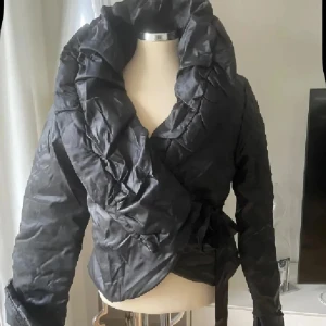 Svart omlott pufferjacka med knytband - Snygg svart pufferjacka med stor krage och omlottmodell. Jackan har ett glansigt tyg och knyts i sidan med ett brett satinband. Långärmad och croppad passform som ger en cool siluett. Perfekt för dig som vill sticka ut med en unik jacka. Sista bilden visar lite ljusare streck , ej fläckar. Jackan är oanvänd 