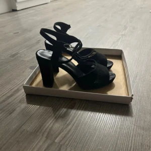 Svarta sandaler med klack och mockadetaljer - Snygga svarta sandaler med bred klack och öppen tå. Remmarna är i mocka och har spänne vid ankeln för extra support. Perfekta för dig som vill ha en stilren och trendig look med lite höjd.