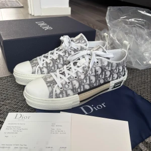 Dior B23 sneakers i vit och grå - Säljer ett par Dior B23 sneakers med ikoniskt monogrammönster i vit och grå ton. Skorna har snörning, platt sula och rund tå. Materialet är en mix av syntet och skinn, med detaljer i silver och en transparent finish på ovandelen. Snygg logotyp på sidan och innersulan.