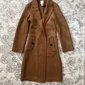 Brun kappa från H&M i ullmix XS - Snygg brun kappa från H&M i klassisk modell med långa ärmar, bred krage och två fickor med svarta knappar. Jackan är rak i modellen och tillverkad i en mjuk ullmix som passar perfekt för höst och vinter.