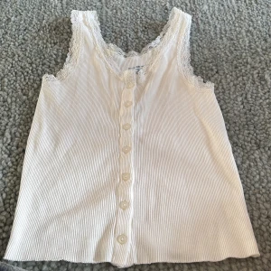 Vit ribbad linnetopp från Brandy Melville - Supersöt vit ribbad linnetopp från Brandy Melville med spetskant runt halsringningen och knäppning framtill. Toppens material är mjuk bomull och den är ärmlös med en feminin och fräsch look. Perfekt att styla med jeans eller kjol för en chill vibe.