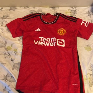 Manchester United matchtröja Adidas S - Röd Manchester United fotbollströja från Adidas med svarta detaljer och vita logotyper. Klassisk klubbmärke på bröstet, TeamViewer-tryck och DXC Technology på ärmen. Kortärmad modell i lätt funktionsmaterial, perfekt för match eller träning.