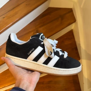 Adidas Campus svarta sneakers mocka - Svarta Adidas Campus sneakers i mocka med klassiska vita ränder på sidorna och vit sula. Skorna har rund tå, platt sula och snörning. Perfekta för dig som gillar en clean och tidlös streetstil.