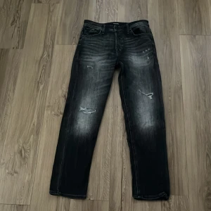Svarta jeans från Jack & Jones - Säljer ett par svarta jeans från Jack & Jones med slitna detaljer och tvättad look. Jeansen har raka ben, klassisk femficksdesign och är tillverkade i denim. Perfekta för dig som gillar en lite ruffare stil.