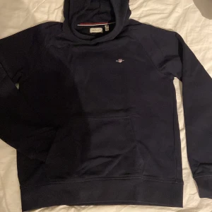 Mörkblå hoodie från GANT - Säljer en stilren mörkblå hoodie från GANT med broderad logga. Tröjan har huva, magficka och långa ärmar. Perfekt för dig som gillar klassisk och enkel stil. Passar runt 170cm