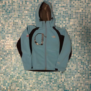 Blå Gore-Tex vindjacka från The North Face - Snygg ljusblå vindjacka från The North Face med svarta detaljer. Jackan har huva, dragkedja framtill, bröstficka med dragkedja och är tillverkad i slitstarkt Gore-Tex-material. Perfekt för blåsiga dagar och outdoor-äventyr.med några märken fråga om bild