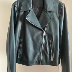 Grön skinnjacka från Massimo Dutti - Snygg mörkgrön skinnjacka från Massimo Dutti med klassisk bikerstil. Jackan har silvriga dragkedjor, bred krage och två fickor framtill. Perfekt för dig som vill ha en stilren och edgy look. Långärmad och med coola detaljer vid midjan.