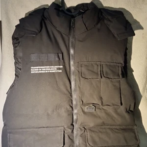 Svart dunväst från Black Squad - Svart dunväst från Black Squad med flera praktiska fickor framtill och coolt tryck på bröstet. Västen har hög krage, grov dragkedja och en urban look. Perfekt för lager-på-lager och streetwear. Transformation Collection-detaljer på vänster sida.