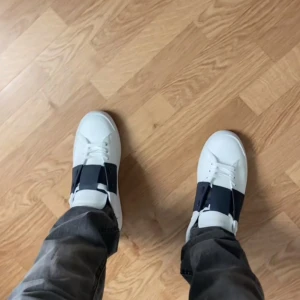 Valentino Garavani Open sneakers vit/svart - Säljer ett par Valentino Garavani Open sneakers i vitt skinn med bred svart läderdetalj över snörningen. Snygg och modern design med platt sula och rund tå. Ikonisk logga på plösen och mönstrad undersida. Perfekt för dig som gillar exklusiva sneakers.