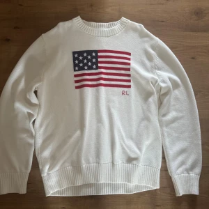 Vit stickad tröja från Ralph Lauren - Snygg vit stickad tröja från Ralph Lauren med amerikansk flagga på bröstet och RL-broderi. Tröjan har rund halsringning, ribbade muddar och är långärmad. Perfekt för dig som gillar klassisk och stilren design.