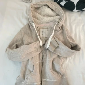 Beige hoodie med teddyfoder från H&M - Mysig beige hoodie från H&M L.O.G.G. med fluffigt teddyfoder i hela luvan och insidan. Tröjan har dragkedja, stora fickor framtill och vita snören i luvan. Storlek M men skulle säga den är mer S