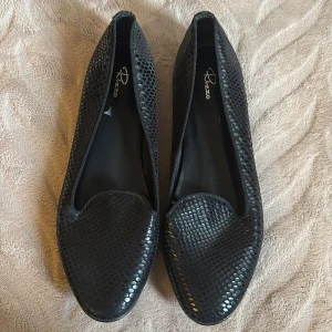 Svarta loafers i ormskinnsmönster från Rizzo - Snygga svarta loafers från Rizzo med ett stilrent ormskinnsmönster. Skorna har rund tå och är tillverkade i skinn, oanvända 