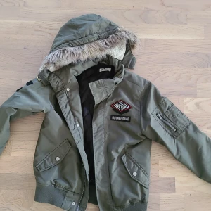 Grön bomberjacka med fuskpäls från H&M - Snygg militärgrön bomberjacka från H&M med huva och fuskpäls, coola patches och dragkedja på ärmen. Jackan har ribbade muddar, två fickor med tryckknappar och quiltat innerfoder. Perfekt för dig som gillar streetstyle och detaljer.