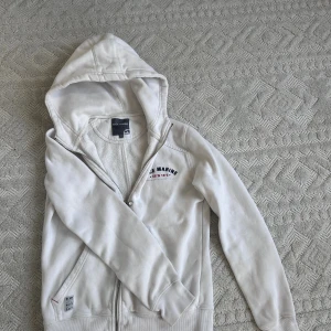 Vit hoodie från Race Marine - Vit hoodie från Race Marine med dragkedja framtill och broderad logga på bröstet. Tröjan har huva, två fickor och ribbade muddar vid ärmslut och nederkant.