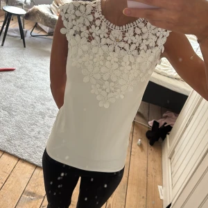 Vit blus med spets och blommor - Supersöt vit blus med ärmlös design och vackert virkad spets upptill. Blusen har blommiga detaljer över bröstet och axlarna som ger en romantisk känsla. Perfekt för dig som gillar feminina och stilrena plagg. Lite nopprig på baksidsn. Ca strlk xs/s