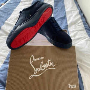 Mörkblå sneakers från Christian Louboutin - Snygga mörkblå sneakers från Christian Louboutin i mocka med klassisk röd sula. Skorna har rund tå, platt sula och snörning. Insidan är fodrad i beige läder för extra komfort. Perfekta för dig som vill ha en lyxig och stilren look.