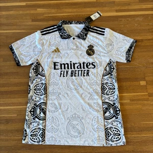 Real Madrid vit matchtröja Adidas M  | Klassisk design - Säljer en Real Madrid fotbollströja från Adidas i vitt med svarta och guldiga detaljer. Tröjan har korta ärmar, svartvit mönstrad krage och klassiska Adidas-ränder på axlarna. Klubbmärket är broderat på bröstet, och materialet är lätt och ventilerande – perfekt för träning, match eller streetwear.  ✔️ Lätt och andas ✔️ Regular fit, korta ärmar ✔️ Aldrig använd  Storlek: M Skick: Ny / aldrig använd 