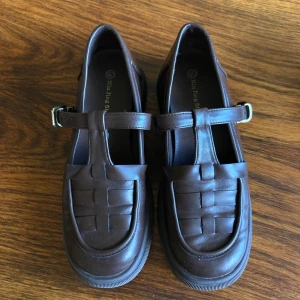 Bruna retro loafers med spänne - Snygga bruna Mary Jane-inspirerade loafers med flätad detalj framtill och justerbart spänne i metall. Skorna har rund tå, platt sula och är gjorda i skinnimitation. Storlek 39, men små i storleken. Knappt används så superfint skick. Perfekta för dig som gillar retrovibbar och chunky stil. Säljer för 300 kr + frakt (köpare står för frakt) 💕