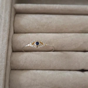 Guldig ring med blå sten och ögonmotiv 17mm - Unik och tunn ring i guldton med ett ögonformat motiv framtill. Ringen har en blå sten i mitten och små klara stenar runtom som ger extra bling. Perfekt för dig som gillar smycken med symbolik och vill sticka ut med en cool detalj.