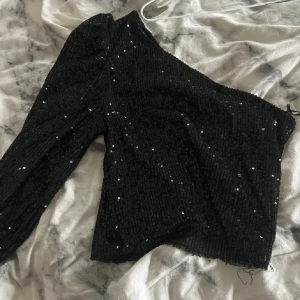 Svart paljett topp från Bik Bok - Säljer en svart oneshoulder topp från Bik Bok, täckt med glittriga paljetter. Toppen har lång ärm på ena sidan och är figurnära i modellen. Perfekt för att sticka ut med en cool och festlig vibe. Materialet är mjukt och har en snygg glans.