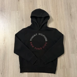 Svart hoodie Tommy Hilfiger NYC S - Svart hoodie från Tommy Hilfiger i storlek S med tryck i vitt och rött på bröstet där det står 'Tommy Hilfiger New York City' i en cirkel. Klassisk huva och ribbade muddar. Perfekt för en chill och stilren look.