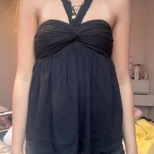 Svart halterneck topp med twist - Säljer en svart halterneck topp med twistad detalj framtill och lös passform nedtill. Toppen har breda axelband som korsas i nacken och är tillverkad i ett tunt, flowy tyg som ger en snygg siluett. Perfekt för dig som gillar trendiga och stilrena plagg.