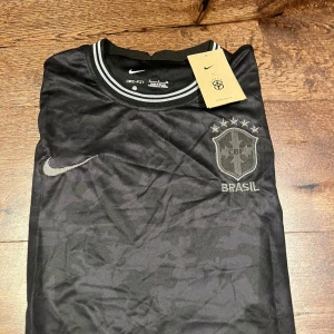 Svart Brasil Nike fotbollströja S - Snygg svart Brasilien fotbollströja från Nike med diskret camomönster och broderad CBF-logga på bröstet. Tröjan har korta ärmar, rund hals med gråa ränder och en stor drake tryckt på ryggen. Tillverkad i Dri-FIT material som andas.