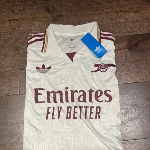 Arsenal x Adidas vit fotbollströja S - Snygg Arsenal fotbollströja från Adidas i vitt med vinröda detaljer och guldinslag. Klassisk krage, broderade loggor och diskret geometriskt mönster i tyget. Tillverkad i lätt och ventilerande material.