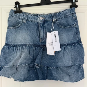 Volangkjol i denim från Gina Tricot - Säljer en blå jeanskjol från Gina Tricot med volangdetaljer och råa kanter. Kjolen har klassisk femficksdesign, bälteshällor och knäppning fram. Perfekt för dig som gillar en trendig och avslappnad stil. Aldrig använd!