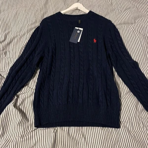 Mörkblå stickad tröja Polo Ralph Lauren - Kabelstickad mörkblå tröja från Polo Ralph Lauren med klassisk röd broderad logga på bröstet. Perfekt för hösten eller nu till vintern. .