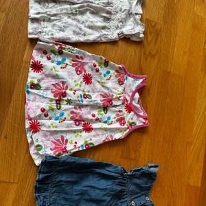 Tre söta sommarklänningar i olika stilar - Tre barnklänningar: en vit med lila blommönster och spetsdetaljer, en färgglad blommig klänning med rosa kant och ärmlös design, samt en jeansklänning med knappar framtill och volang vid axeln. Perfekta för varma dagar och lek.