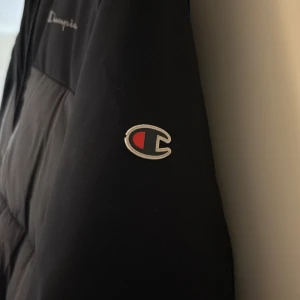 Svart pufferjacka från Champion  - Svart pufferjacka från Champion i storlek 13/14y. Jackan har huva, hel dragkedja och två fickor framtill. Endast använd en vinter och är i mycket bra skick med med inga defekter. Säljer för att jag inte använder längre. Pris kan diskuteras 