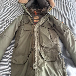 Grön parkas från Parajumpers med päls - Säljer en grön parkas från Parajumpers Masterpiece Series. Jackan har flera stora fickor, gul detaljrem, päls på huvan och mysigt foder. Klassisk patch på ärmen och dragkedja med knappar. Perfekt för kalla dagar och riktigt snygg streetstil. Huvudragkedja är utbytt.